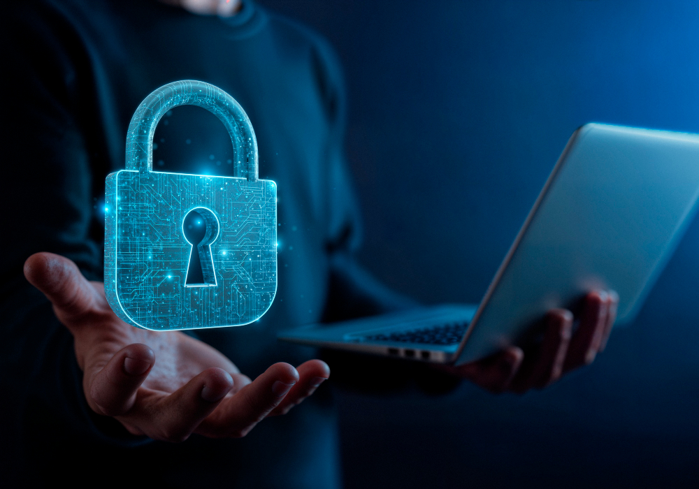Osoba trzyma otwarty laptop, a nad jej wyciągniętą dłonią unosi się świetlista, cyfrowa kłódka symbolizująca ochronę danych i cyberbezpieczeństwo.