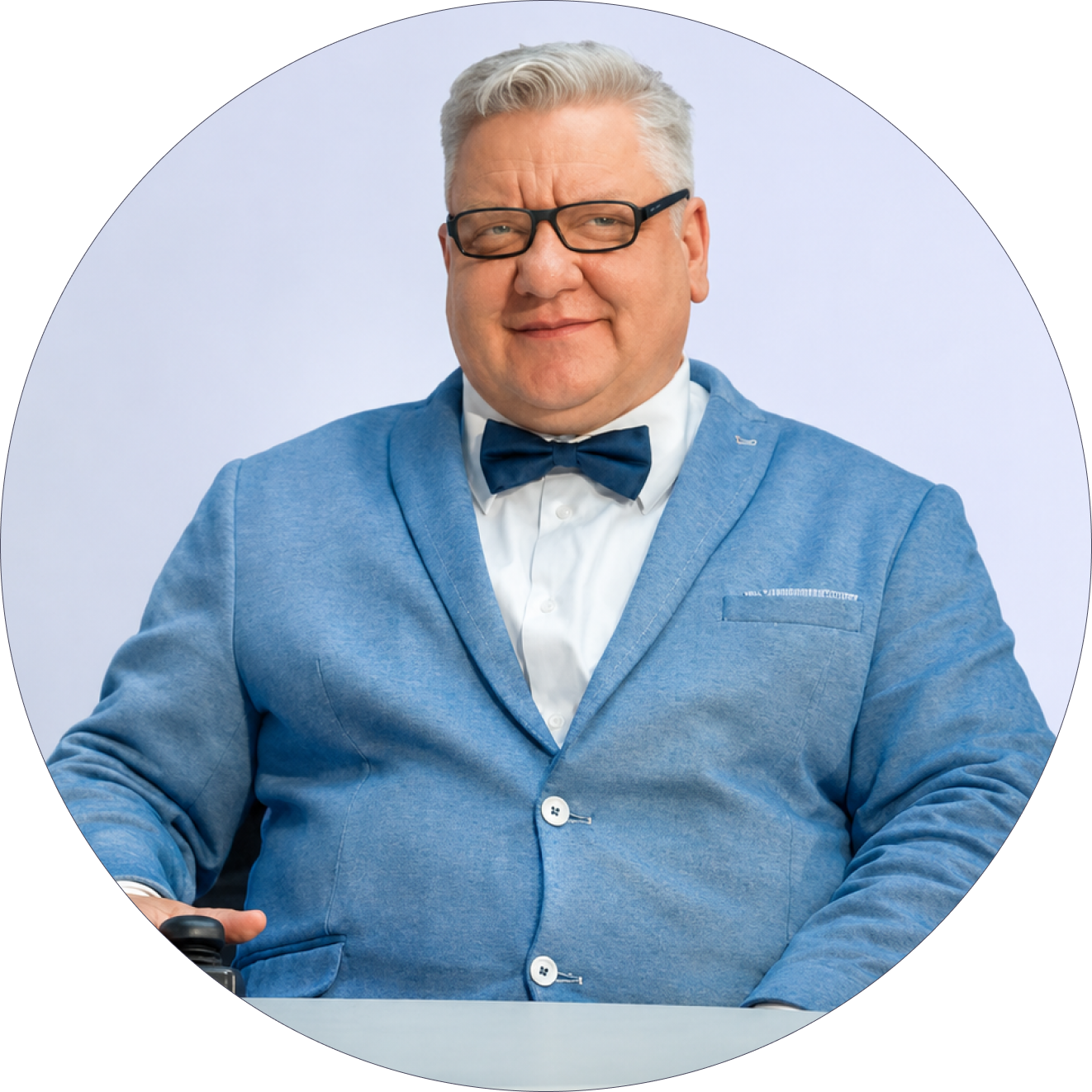 Andrzej Byszewski siedzi na elektrycznym wózku za okrągłym stołem. Ma na sobie jasnoniebieską marynarkę, białą koszulę, granatową muszkę i okulary. Lekko się uśmiecha na jasnym, jednolitym tle.