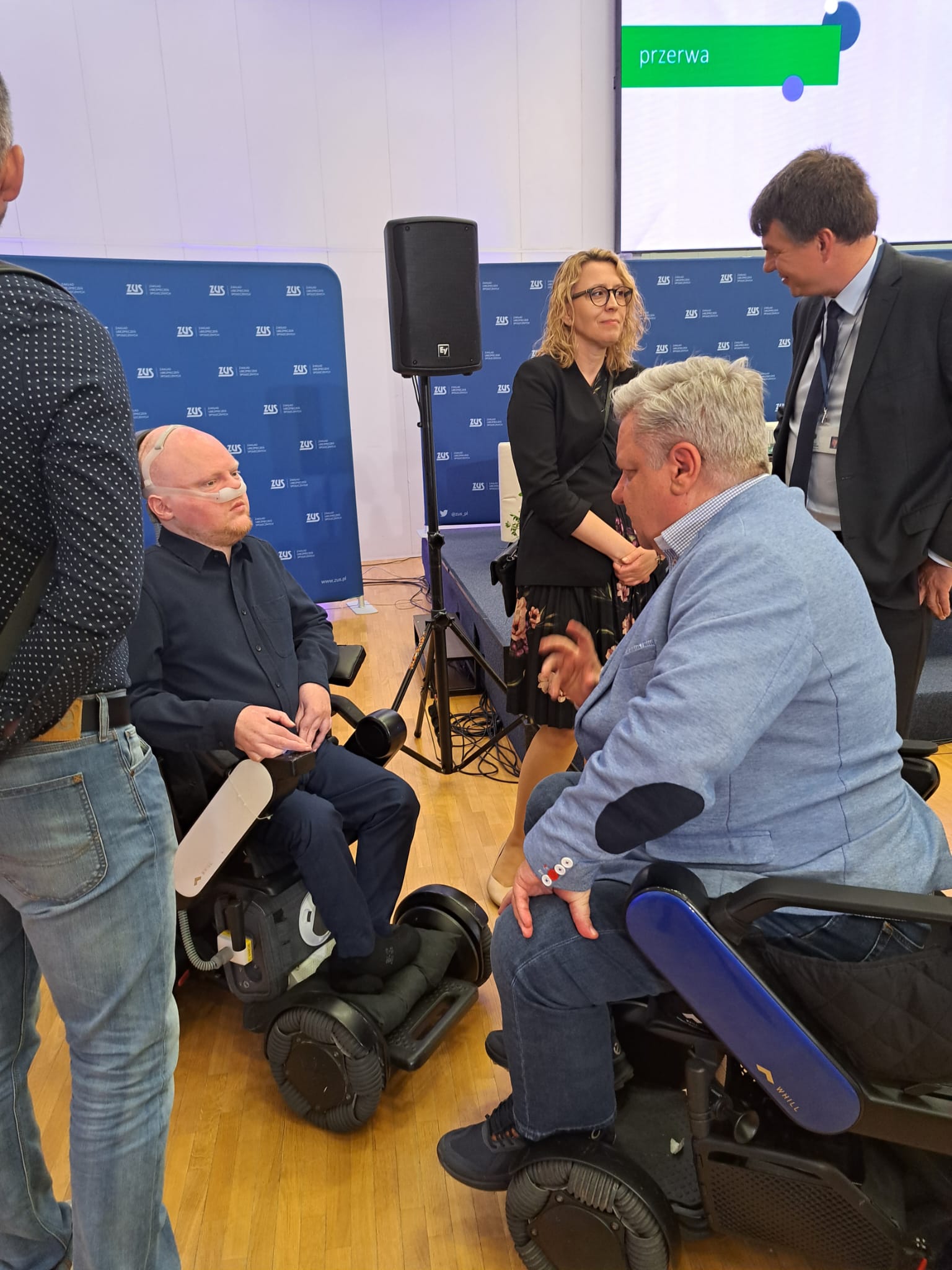 Mężczyźni na wózkach elektrycznych rozmawiają z innymi uczestnikami wydarzenia przy stanowisku konferencyjnym.